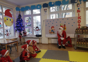 Widok na salę przedszkolną, czworo dzieci siedzących na dywanie oraz Świętego Mikołaja. W tle świąteczno- zimowe dekoracje.