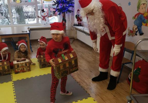 Widok na salę przedszkolną, dzieci oraz Świętego Mikołaja i dziewczynkę, która odbiera od niego prezent.