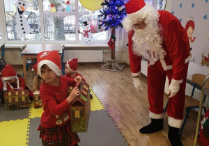 Widok na salę przedszkolną, dzieci oraz Świętego Mikołaja i dziewczynkę, która odbiera od niego prezent.