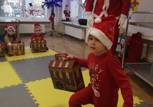 Widok na salę przedszkolną, dzieci oraz Świętego Mikołaja i chłopca, który odbiera od niego prezent.