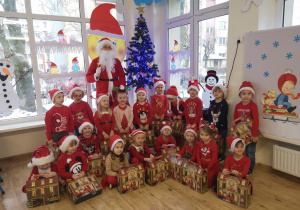 Maluchy w czerwonych strojach, z prezentami pozują do zdjęcia grupowego ze Świętym Mikołajem.