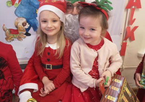 Widok na dwie dziewczynki, które pozują do zdjęcia siedząc na kolanach Świętego Mikołaja. W tle tablica z obrazkiem chłopca niosącego prezenty.