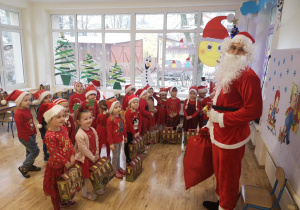Widok na grupę przedszkolaków w mikołajkowych strojach. Dzieci stoją z prezentami przed Świętym Mikołajem. W tle świątecznie udekorowane okno, za którym widać ośnieżone drzewo i krzewy.