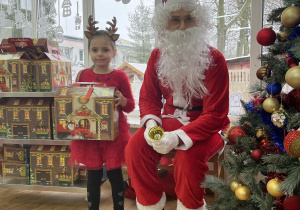 Dziewczynka pozuje do zdjęcia ze Świętym Mikołajem trzymając w rękach prezent.