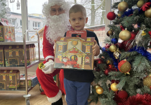 Chłopiec pozuje do zdjęcia ze Świętym Mikołajem trzymając w rękach prezent.
