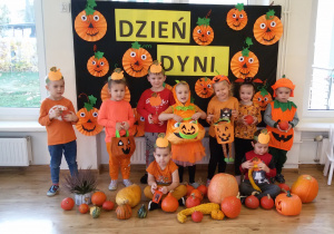 Widok na dzieci w pomarańczowych strojach, które pozują do zdjęcia grupowego, na tle tablicy z napisem: Dzień Dyni.