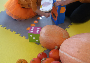 Widok na stertę dyni, dużą, kolorową kostkę do gry oraz pudełko z żarłoczną dynią, do którego Amelka wrzuca pomarańczowe papierowe kółeczka.