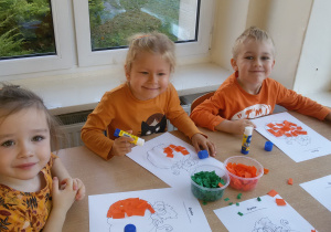 Widok na 3 dzieci, które wyklejają obrazek przedstawiający dynię. Na stoliku stoją pojemniki z bibułką w kolorze zielonym i pomarańczowym.