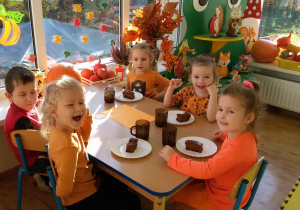 Widok na siedzące przy stoliku dzieci, przed którymi stoją kubeczki oraz talerzyki z ciastem dyniowym.