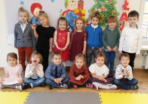 Maluchy w odświętnych strojach pozują do zdjęcia grupowego na tle bożonarodzeniowej dekoracji.