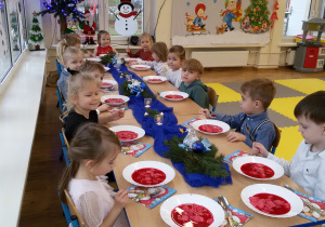 Widok na dzieci siedzące przy odświętnie udekorowanym stole. Przed nimi stoją talerze z czerwonym barszczem