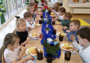 Widok na długi, odświetnie udekorowany stół, przy którym dzieci jedzą wigilijny posiłek.