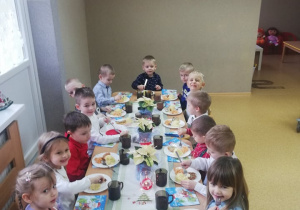 Dzieci ubrane w odświętne stroje wspólnie przy dużym stole zjadają wigilijny obiad.
