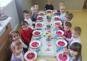 Dzieci ubrane w odświętne stroje wspólnie przy dużym stole zjadają wigilijny barszcz.