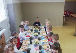 Dzieci ubrane w odświętne stroje wspólnie przy dużym stole zjadają wigilijny obiad.