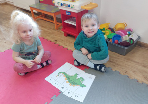 Chłopiec i dziewczynka siedzący na macie prezentują ułożonego dinozaura.