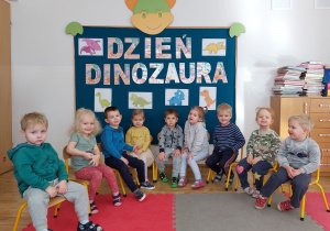 Maluszki pozują do zdjęcia grupowego na tle tablicy z dekoracją z okazji Dnia Dinozaura.