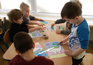 Widok na siedzące przy stoliku dzieci, które stemplują palcami sylwety dinozaurów.