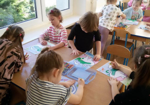 Widok na siedzące przy 2 stolikach dzieci, które stemplują palcami sylwety dinozaurów.