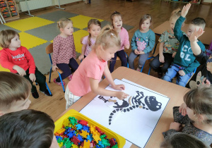 Antosia układa papierową kość na planszy ze szkieletem dinozaura. W tle siedzące na krzesłach dzieci.