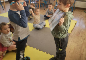 Widok na 2 chłopców, z których jeden trzyma obrazek w 3D, a drugi ogląda go w okularach. W tle widać siedzące na macie dzieci.