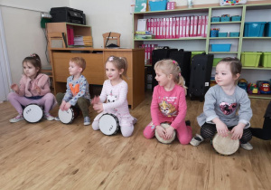 Widok na dzieci grające na bębnach afrykańskich. W tle biurko i szafki z pojemnikami na klocki.