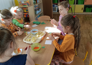 Dziewczynki siedzą przy stoliku i sklejają wodą chrupki kukurydziane w kształt dyni. Na stole taca z chrupkami kukurydzianymi, miseczka z wodą, pędzle i miseczka z farbą pomarańczową.