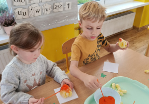 Chłopiec i dziewczynka malują pomarańczową farbą dynię wykonaną z chrupek.