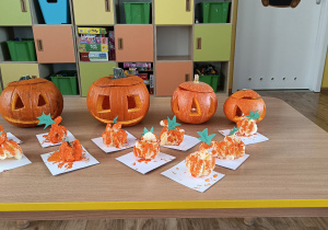 Na stole prezentacja 4 lampionów zrobionych z dyni oraz prac dzieci „dynie z chrupek” w kolorze pomarańczowym.