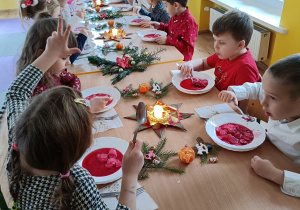 Dzieci siedząc przy stole jedzą potrawę wigilijną (barszcz czerwony). Na stole świąteczna dekoracja (gwiazdy z serwetek, gałązki świerku, świeczki).