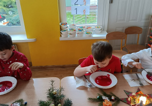 Dzieci siedząc przy stole jedzą potrawę wigilijną (barszcz czerwony). Na stole świąteczna dekoracja (gwiazdy z serwetek, gałązki świerku, świeczki).
