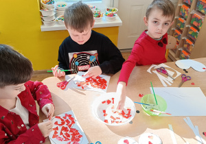 3 chłopców siedzi przy stole i wyklejają papierowe serduszko czerwonymi kawałkami papieru.