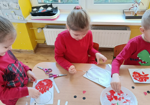 3 dziewczynki siedzą przy stole i wyklejają papierowe serduszko czerwonymi kawałkami papieru.