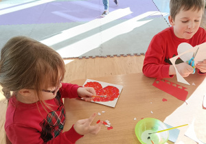 Dwoje dzieci siedzi przy stole. Dziewczynka wykleja serce kawałkami czerwonego papieru, chłopiec wycina serce nożyczkami.