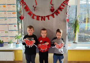 3 chłopców stojąc pozuje do zdjęcia trzymając w ręku papierowe serduszko, w tle napis Walentynki.
