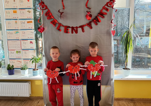 Troje dzieci stojąc pozuje do zdjęcia trzymając w ręku papierowe serduszko, w tle napis Walentynki.