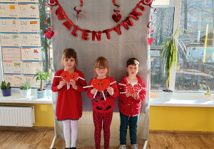 Troje dzieci stojąc pozuje do zdjęcia trzymając w ręku papierowe serduszko, w tle napis Walentynki.