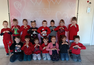 Dzieci pozują do zdjęcia trzymając ręce w kształcie serca. W tle baner z sercami.