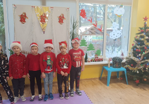 Sześcioro dzieci pozuje do zdjęcia ubrane w czerwone stroje i mikołajkowe czapeczki. W tle tablica udekorowana postaciami Mikołaja oraz ubrana choinka.