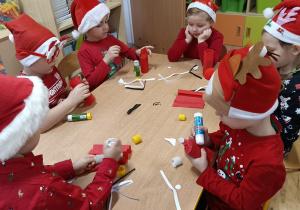 Dzieci siedzą przy stoliku i wykonują postać Mikołaja z rolki papierowej obklejonej czerwonym papierem i elementami: czapeczka, pasek, broda.