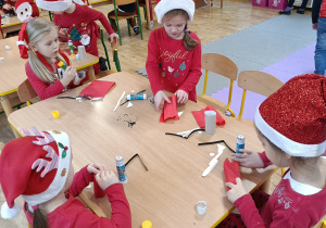 Dzieci siedzą przy stoliku i wykonują postać Mikołaja z rolki papierowej obklejonej czerwonym papierem i elementami: czapeczka, pasek, broda.
