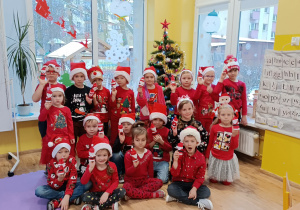 Dzieci pozują do zdjęcia w czerwonych strojach z czapeczkami mikołajkowymi. W rękach trzymają postać Mikołaja wykonanego z rolki papierowej.