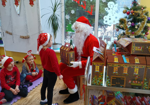 Dziewczynka stoi przed Mikołajem siedzącym na krześle, który trzyma upominek „domek z kartonu”.