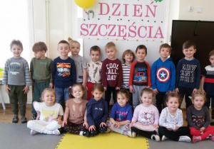 Maluchy pozują do zdjęcia grupowego na tle tablicy z napisem Dzień Szczęścia.