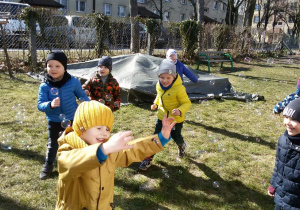 Widok na grupę bawiących się w ogrodzie przedszkolnym dzieci, które chwytają spadające bańki mydlane.