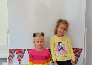 Dziewczynki pozują do zdjęcia. W tle tablica magnetyczna, na której znajduje się kolorowa girlanda papierowa w balony.