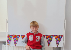 Chłopiec pozuje do zdjęcia. W tle tablica magnetyczna, na której znajduje się kolorowa girlanda papierowa w balony.