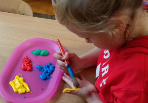 Dziewczynka maluje pędzelkiem swoją dłoń wybranym kolorem farby.