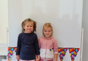Dziewczynki pozują do zdjęcia. W tle tablica magnetyczna, na której znajduje się kolorowa girlanda papierowa w balony.