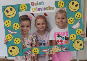 Dziewczynki pozują do zdjęcia stojąc za ramką ozdobioną uśmiechami.